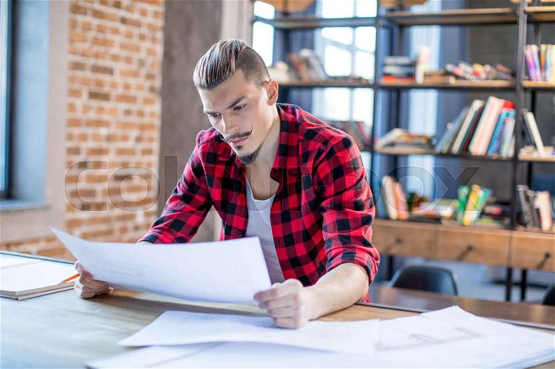 Pensive yound male architect examining blueprints in office , stock photo