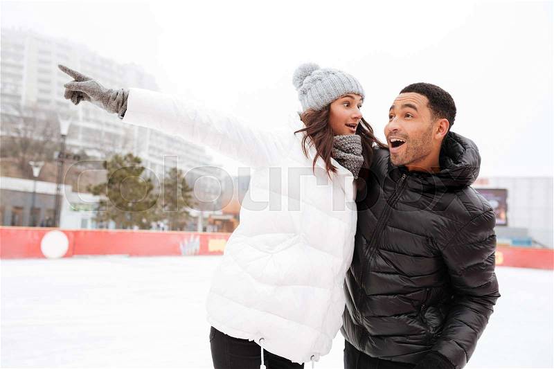 Image of young cheerful loving couple skating at ice rink outdoors. Looking aside and pointing, stock photo