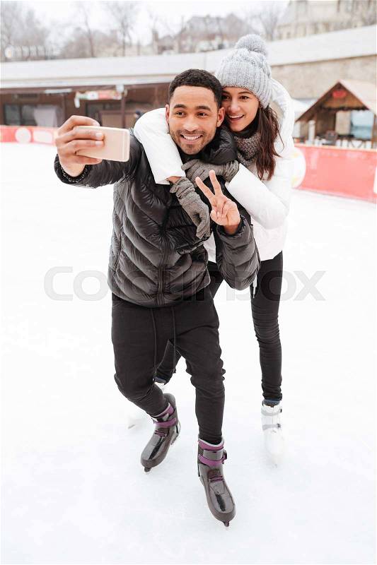 Photo of young cheerful loving couple skating at ice rink outdoors. Make a selfie by phone and make peace gesture, stock photo