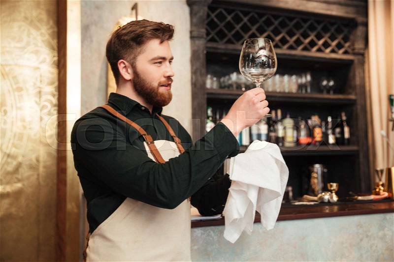 Photo of amazing young bearded man bartender standing in cafe. Looking aside and wipe the glass, stock photo