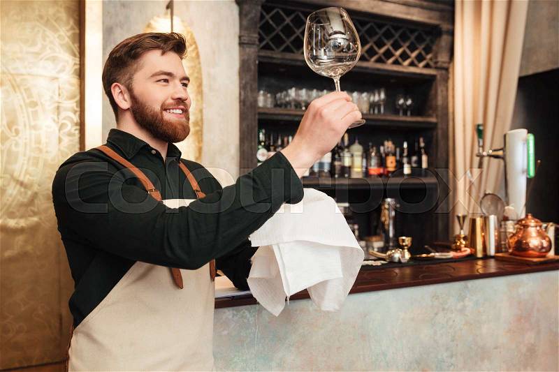 Image of attractive young bearded man bartender standing in cafe. Looking aside and wipe the glass, stock photo