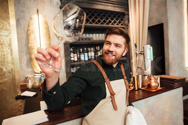 Image of attractive young bearded man bartender standing in cafe. Looking aside and wipe the glass, stock photo