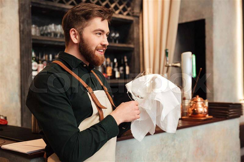 Photo of happy young bearded man bartender standing in cafe. Looking aside and wipe the glass, stock photo