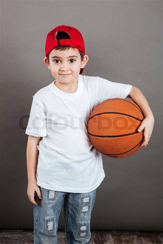Happy little boy in cap standing and holding basketball ball, stock photo
