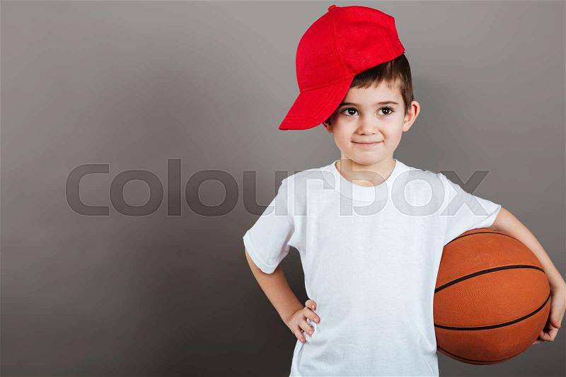 Cute little boy in red cap holding basketball ball over grey background, stock photo