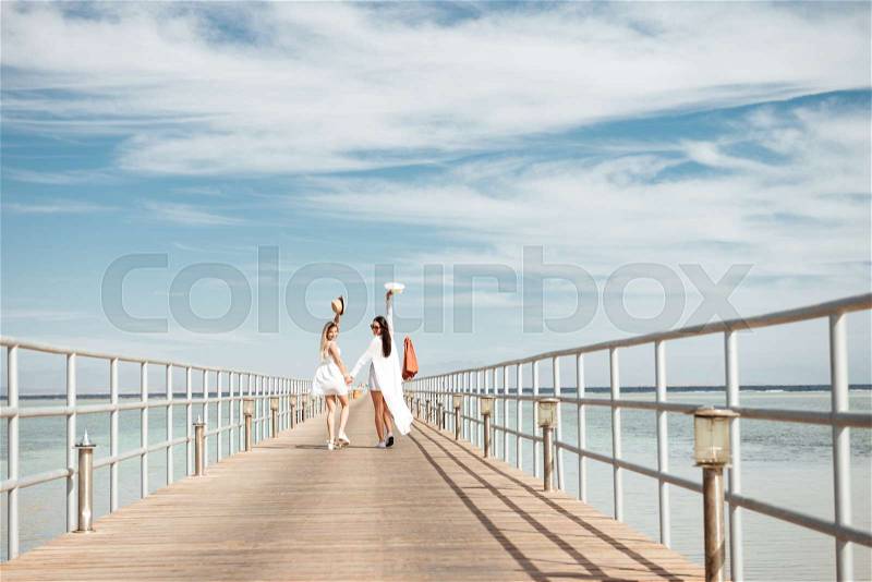 Two happy young women holding hands, walking on pier and looking back, stock photo