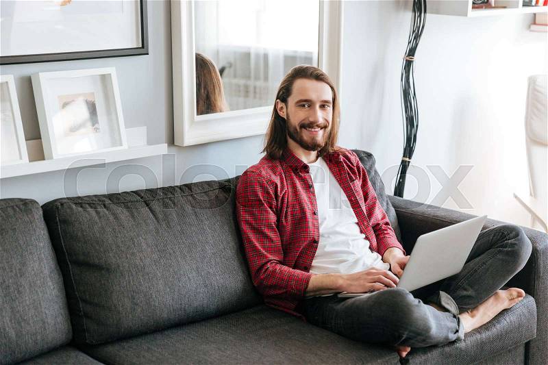 Cheerful bearded young man in plaid shirt using laptop on couch at home, stock photo