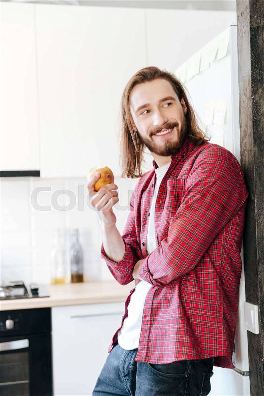 Smiling attractive bearded young man standing and eating apple at home, stock photo