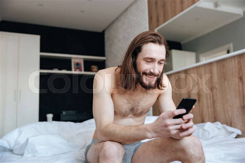 Happy bearded young man using cell phone sitting on bed in bedroom, stock photo