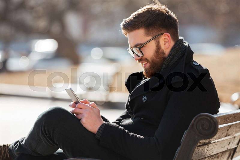 Happy bearded young man in glasses using cell phone in the city, stock photo