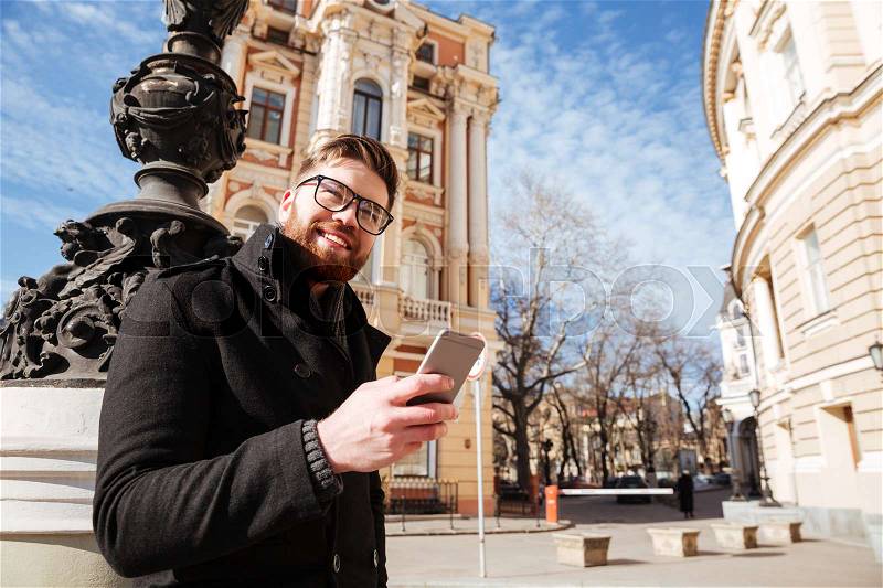Smiling Man in coat and eyeglasses standing on the street and holding phone, stock photo