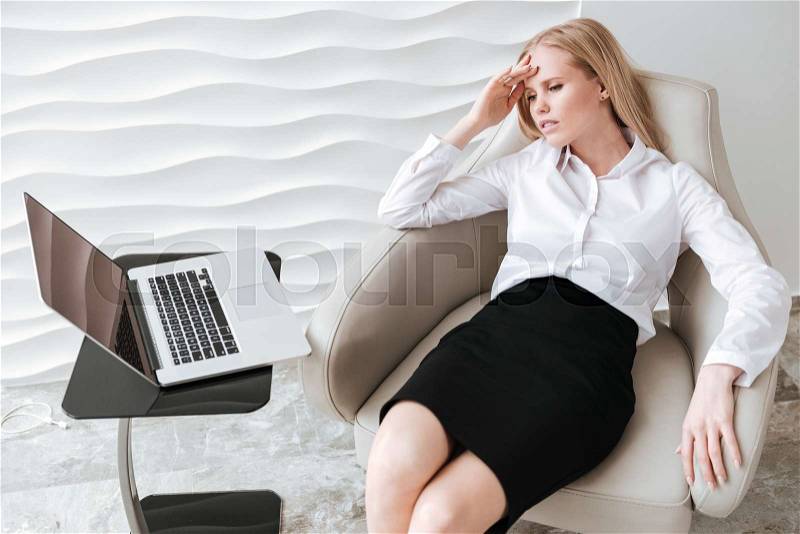 Image of young tired business woman sitting in office on chair. Looking at laptop, stock photo