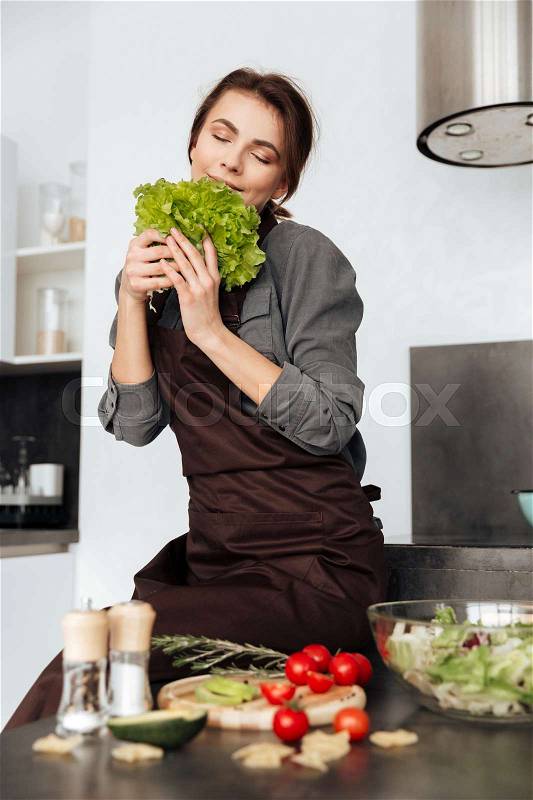 Photo of young woman standing in kitchen smells lettuce leaves and cooking with the tomatoes and avocado, stock photo