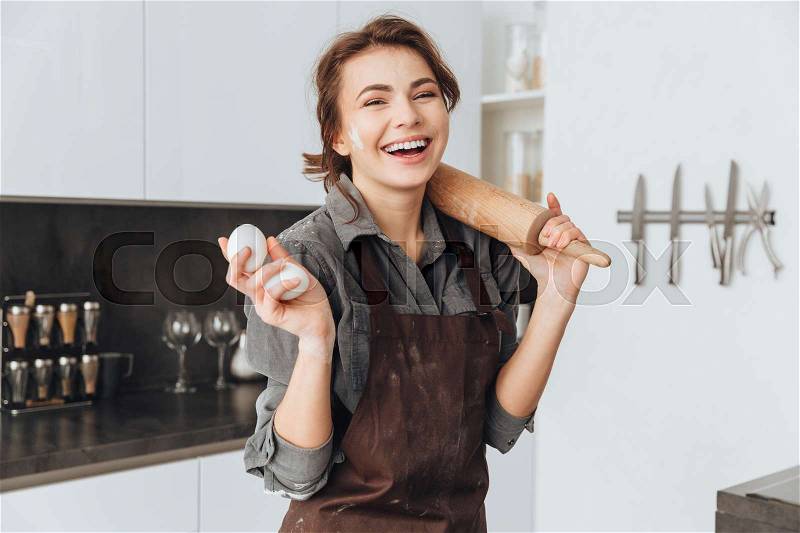 Photo of young laughing lady standing in kitchen and cooking the dough. Looking at camera, stock photo
