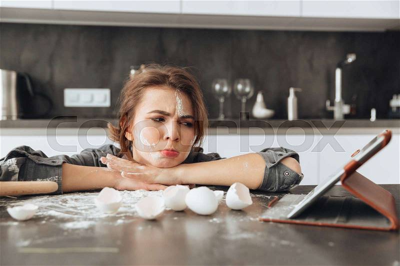 Photo of young sad lady sitting in kitchen and cooking the dough. Looking at tablet computer, stock photo