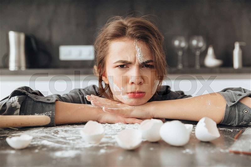 Image of young confused lady sitting in kitchen and cooking the dough. Looking at camera, stock photo