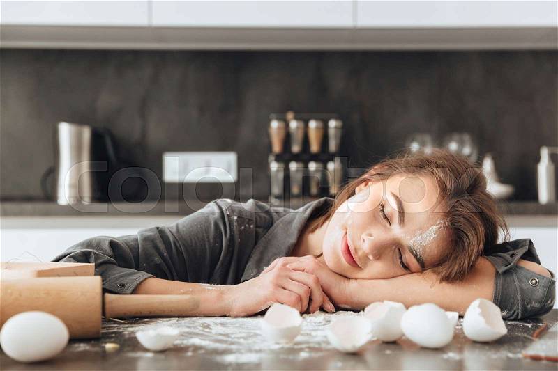 Photo of young cheerful lady sleeping in kitchen after cooking the dough. Eyes closed, stock photo