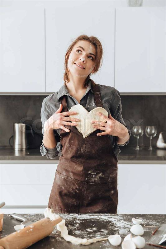 Image of young dreaming lady standing in kitchen while holding the dough heart. Look aside, stock photo