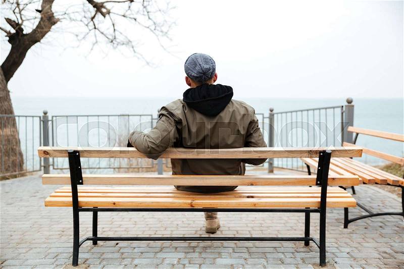Back view picture of young african man wearing hat and scarf sitting on a bench on the street, stock photo