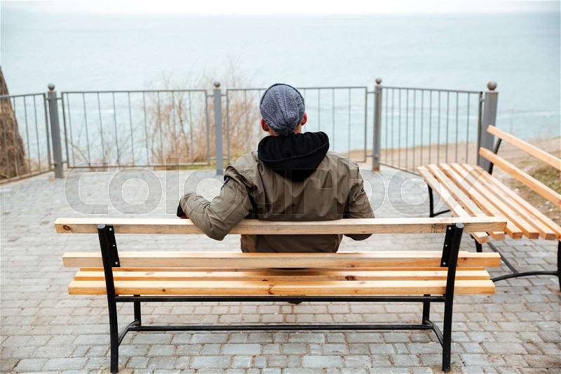 Back view picture of young african man wearing hat and scarf sitting on a bench on the street, stock photo