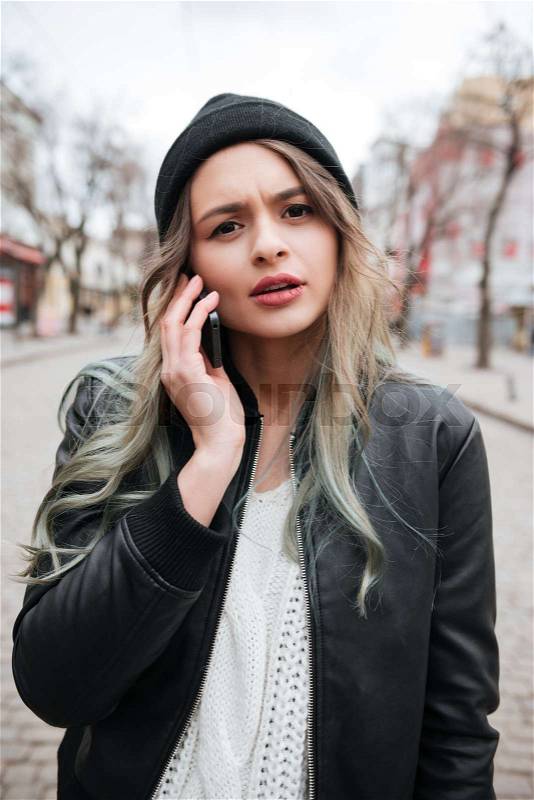 Photo of confused young woman wearing hat walking on the street and talking by mobile phone, stock photo