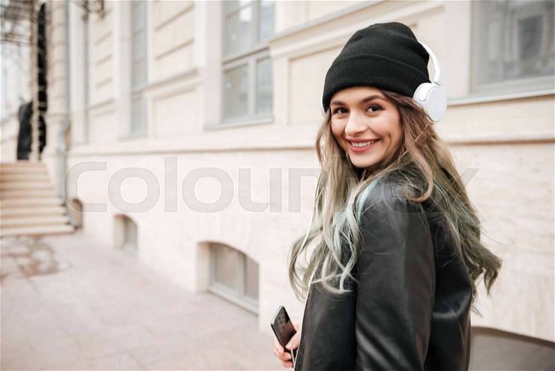 Back view of woman in warm clothes and headphone looking at camera on the street, stock photo