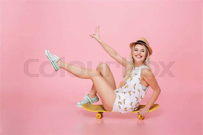Happy charming young woman sitting on skateboard and having fun over pink background, stock photo