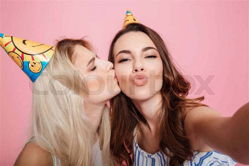 Two happy beautiful young women in birthday hats kissing and talking selfie over pink background, stock photo