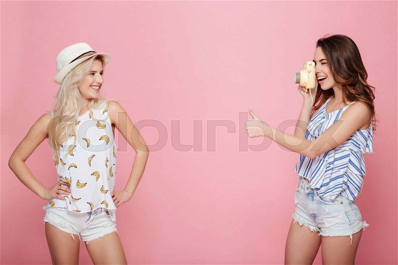 Smiling young woman taking picture of her friend and showing thumbs up over pink background, stock photo