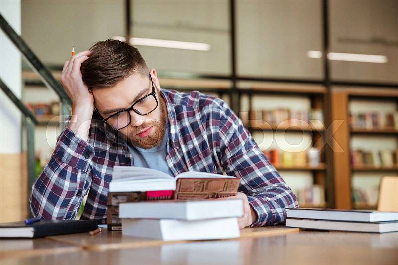 Young guy student reading book with serious face in the library, stock photo