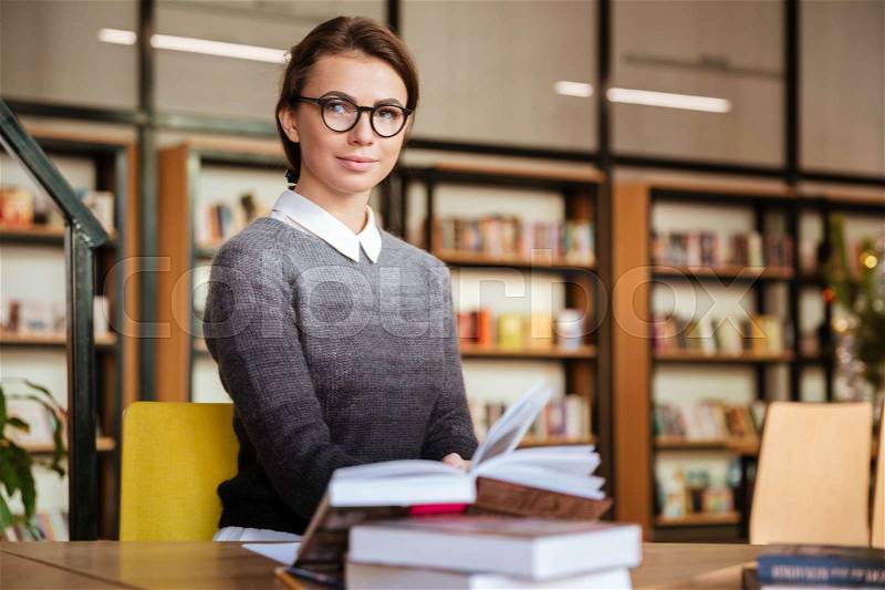 Smiling attractive female student sitting at desk in university library and preparing for exam, stock photo