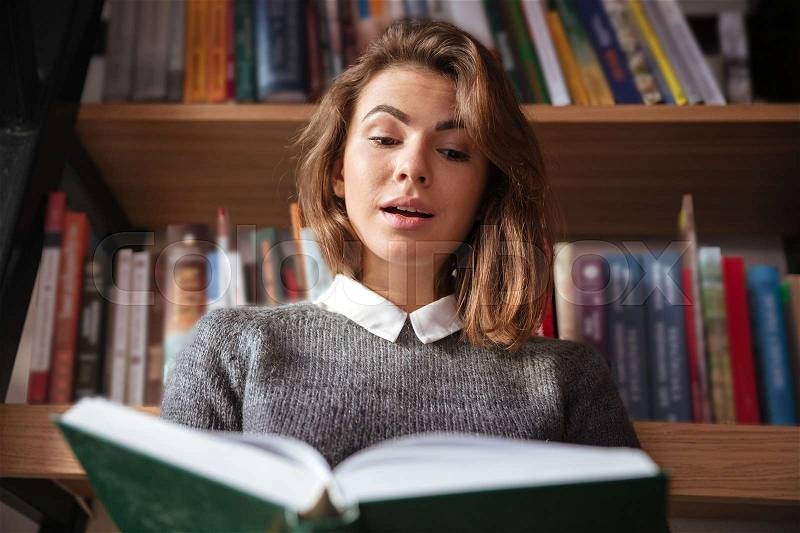 Surprised young woman reading a book at the library, stock photo