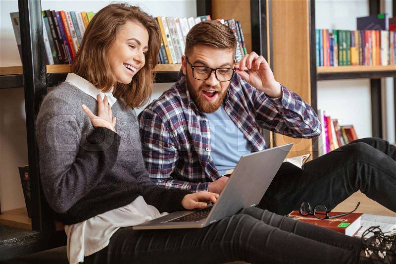 Happy students with laptop computer networking in library while sitting on the floor, stock photo