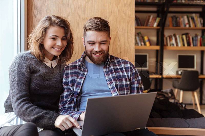 Happy students with laptop computer networking in library while sitting on the windowsill, stock photo