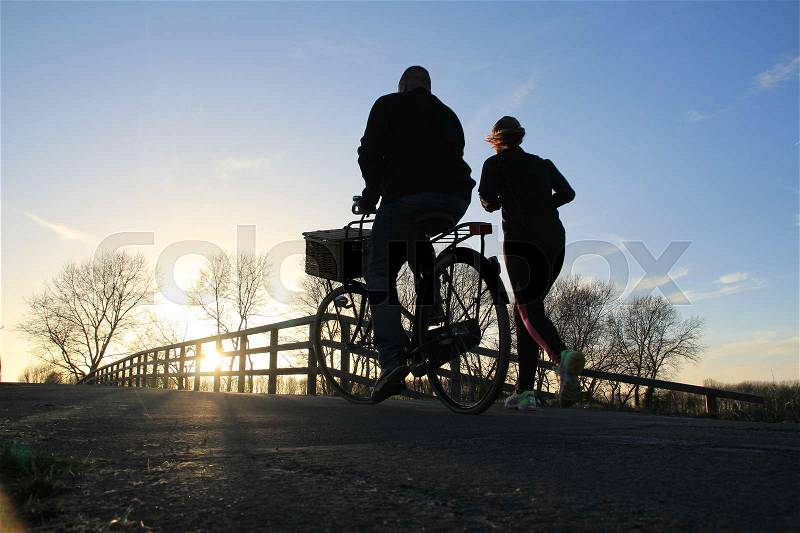 Couple, man is biking and the lady is running over the wooden bridge in the park at the country side at sunset in the soft winter, stock photo