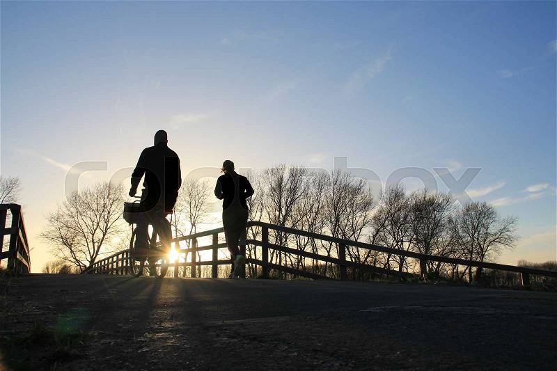 Couple, man is biking and the lady is running over the wooden bridge in the park at the country side at sunset in the soft winter, stock photo