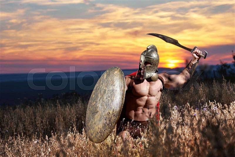 Warrior in helmet with bare torso wearing red cloak like spartan going in attack with iron sword. Incognito male like antique roman soldier holding bronze shield in hand. Sky at sunset, stock photo