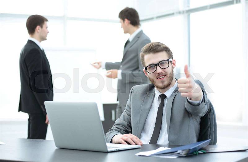 In the foreground is a successful Manager sits at a Desk before an open laptop and showing thumbs up.in the background - colleagues discussing a new presentation . in the photo there is an empty space for your text, stock photo