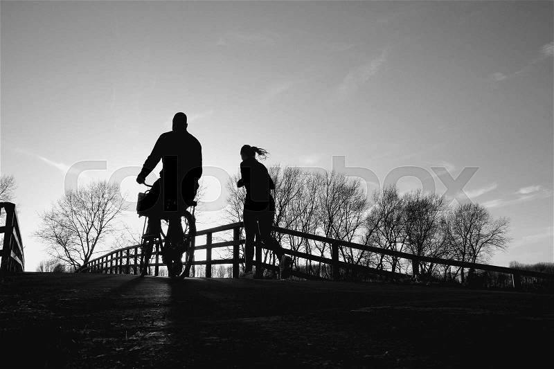 Couple, man is biking and the lady is running over the wooden bridge in the park at the country side at sunset in the soft winter in black and white, stock photo
