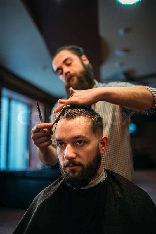 Bearded hairdresser making male hipster hairstyle. Client man in black salon cape visit barbershop, stock photo