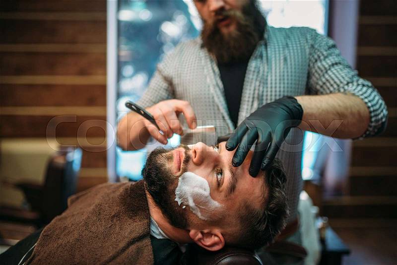 Barber shaves the beard of the client by straight razor at barbershop, stock photo