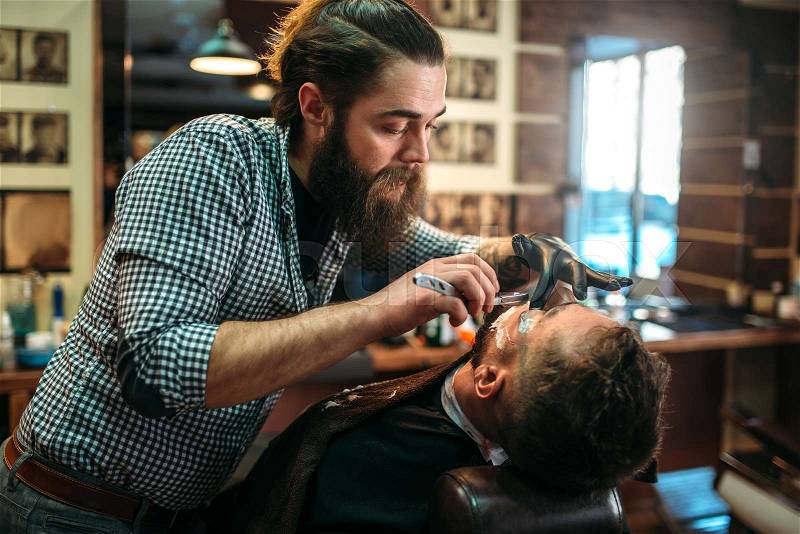 Barber shaves the beard of the client by shaving blade at barbershop. , stock photo