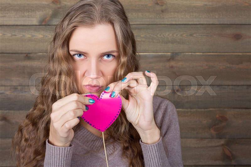 Young female with funny pink heart on a stick, wooden background. Fun photo props and accessories for shoots, stock photo
