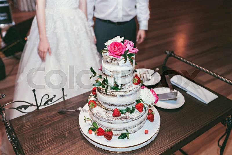 Wedding high cake, decorated with red roses and fresh strawberry, on small table, stock photo