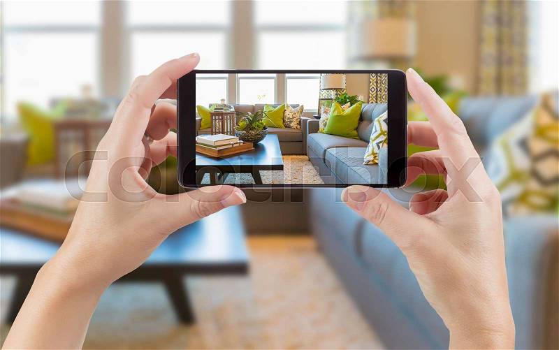 Female Hands Holding Smart Phone Displaying Photo of House Interior Living Room Behind, stock photo