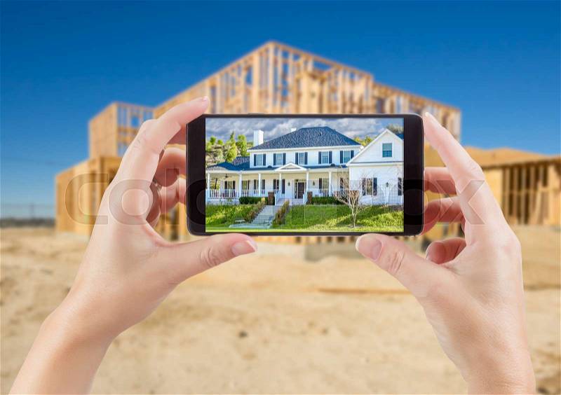 Female Hands Holding Smart Phone Displaying Photo of House with Unfinished Home Construction Behind, stock photo
