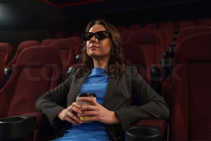 Young woman watches movie 3D at the cinema and drinks coffee, looking at the screen, alone in an empty room, overall plan, stock photo