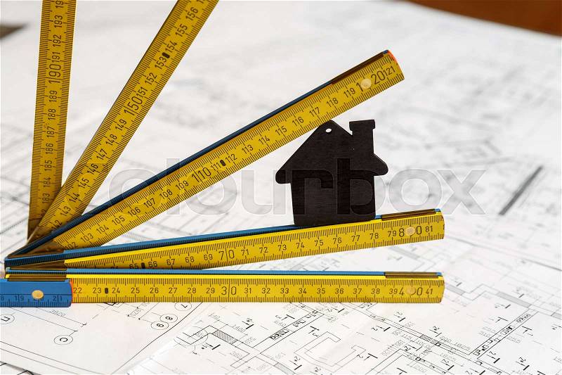 Image of small black toy house and a collapsible yellow and blue ruler standing on top of architecture blueprint plan, stock photo