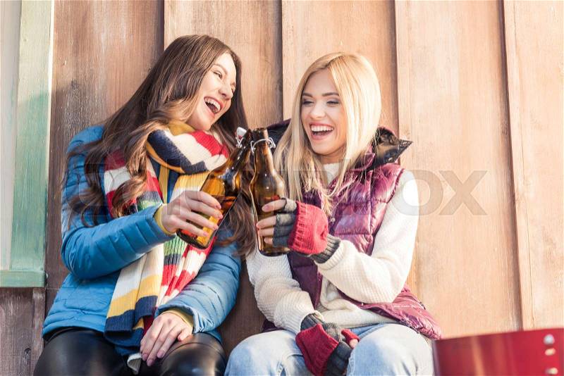 Low angle view of laughing women clinking beer bottles on porch, stock photo