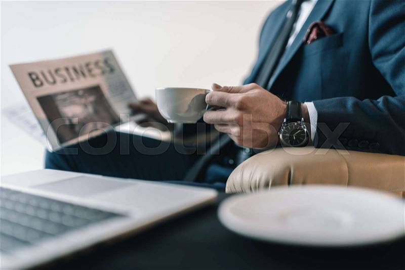 Partial view of businessman with cup of coffee reading newspaper in office, stock photo